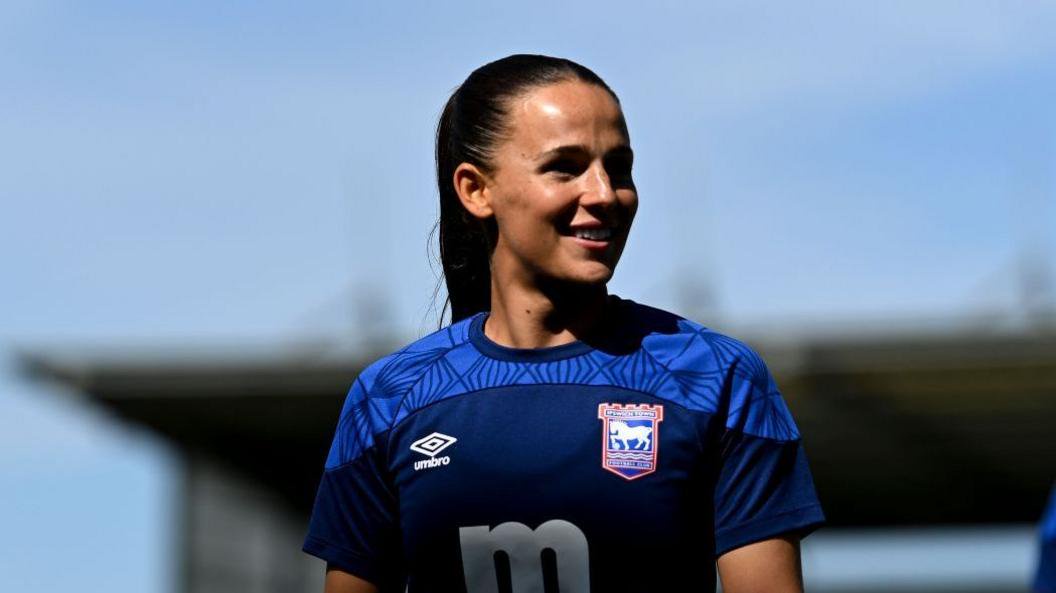 Maria Boswell walking on to the pitch looking to her left. She is wearing blue Ipswich Town kit.