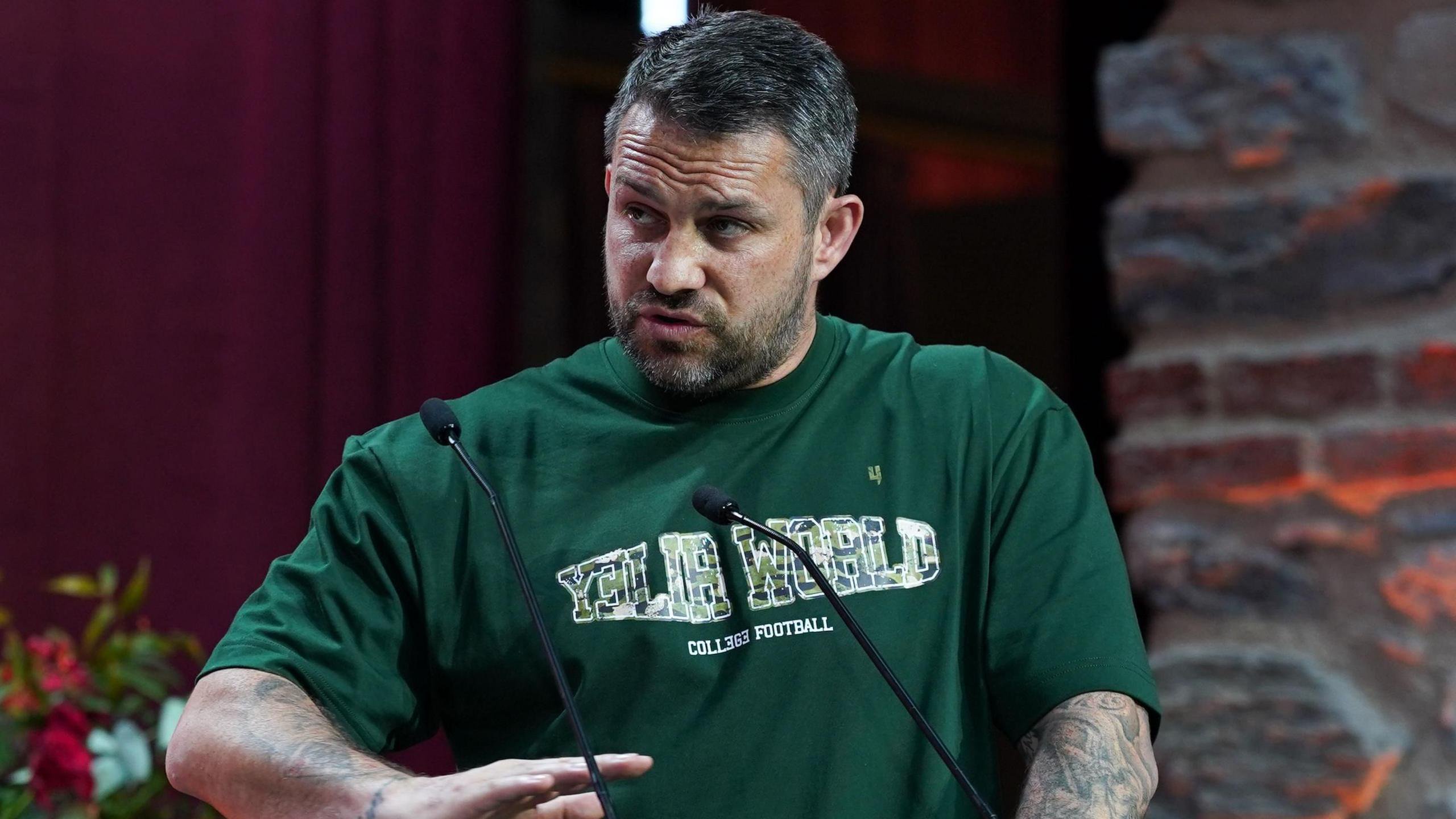 Paul Smith stands behind a grey lectern bearing the International Boxing Association logo, speaking into two microphones. He has short, dark hair, flecked with grey, a beard and tattoos covering parts of both forearms. 
