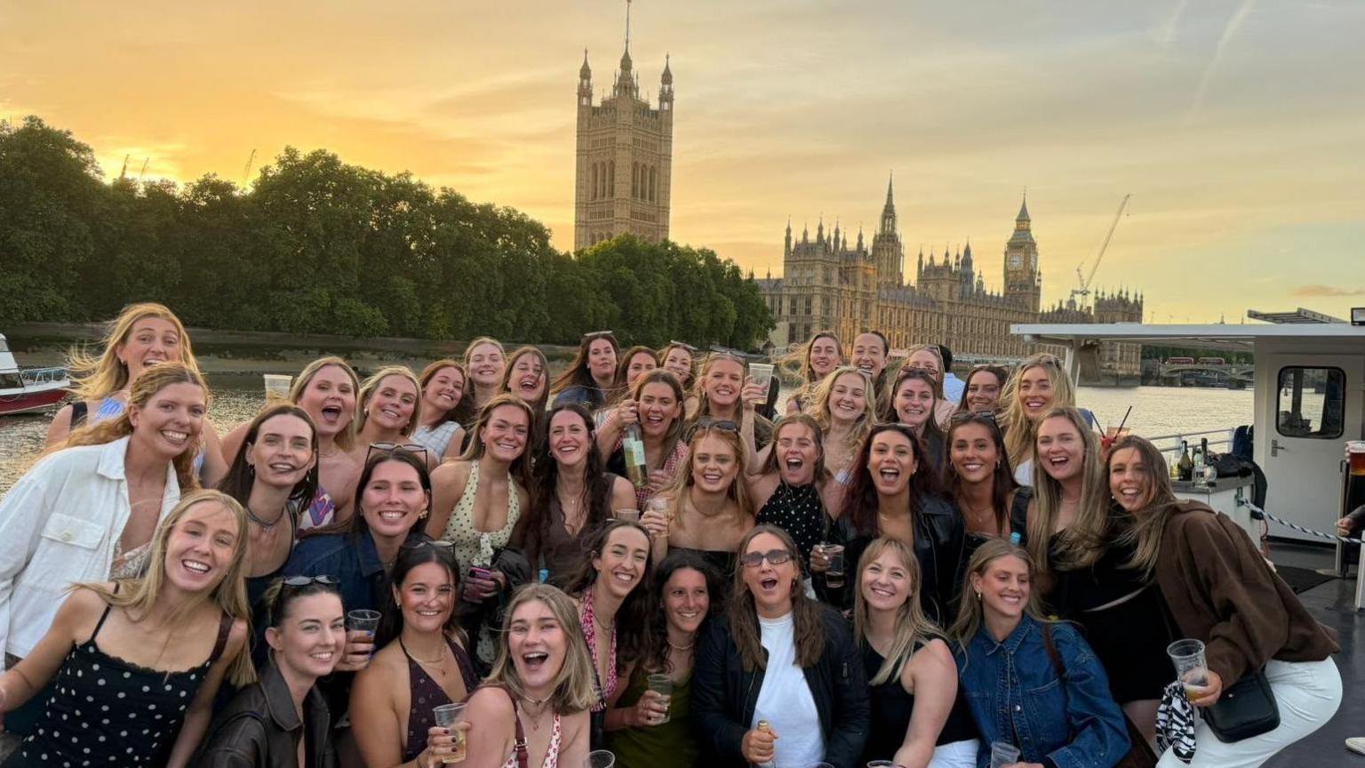 A large group pose for a photograph beside the River Thames at sunset, with the Palace of Westminster and the Elizabeth Tower visible in the background. Many are holding drinks, and a boat is moored alongside the river behind the group.