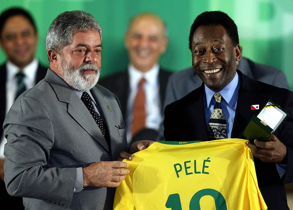 Two men stand together smiling. The man on the left, in a gray suit, holds a yellow jersey labeled "Pelé 10." The man on the right, in a dark suit, also holds a plaque or medal. A blurred crowd is visible in the background.