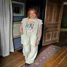 A woman with long red hair, wearing a Def Leppard t-shirt, white vest, and matching wide-leg pants, stands in a room with wooden flooring and framed pictures