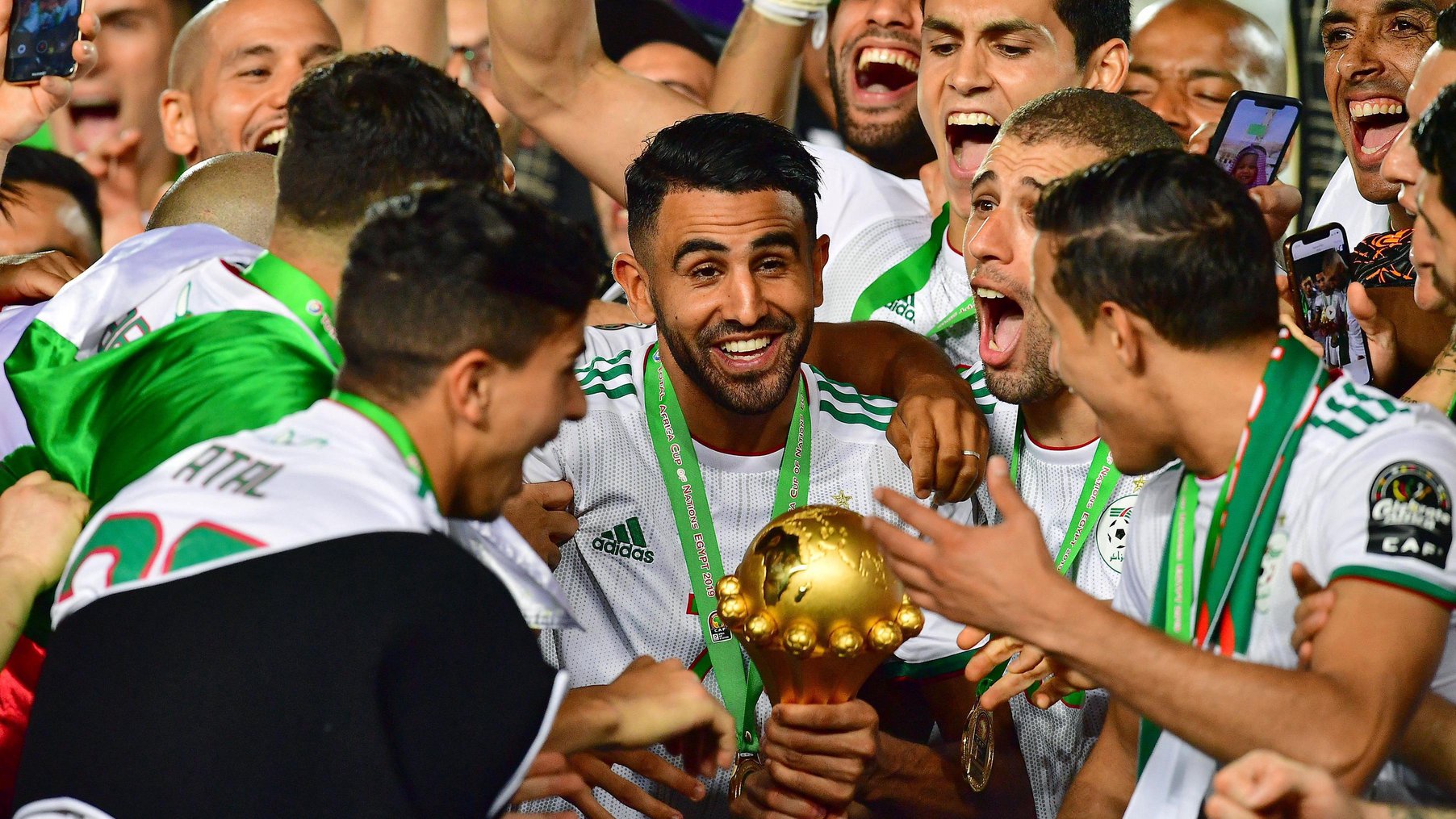Algeria captain Riyad Mahrez, wearing a white shirt and winner's medal on a green ribbon, smiles as he holds the Africa Cup of Nations trophy while surrounded by gleeful team-mates who are celebrating with him after the trophy lift at the 2019 Afcon final
