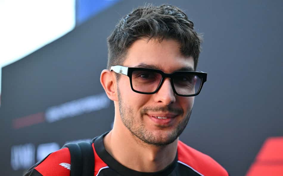Homem com barba por fazer, óculos pretos, camiseta vermelha, sorrindo, em evento de Fórmula 1