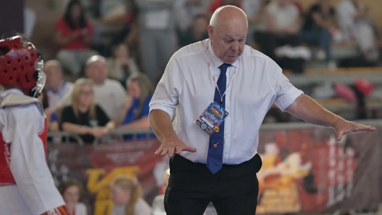 Supplied A martial arts referee in a white shirt, blue tie and black trousers stands in the ring with his arms outstretched, overseeing a match between competitors wearing red, protective headgear and gloves. Spectators watch from the seating behind barriers in the background.