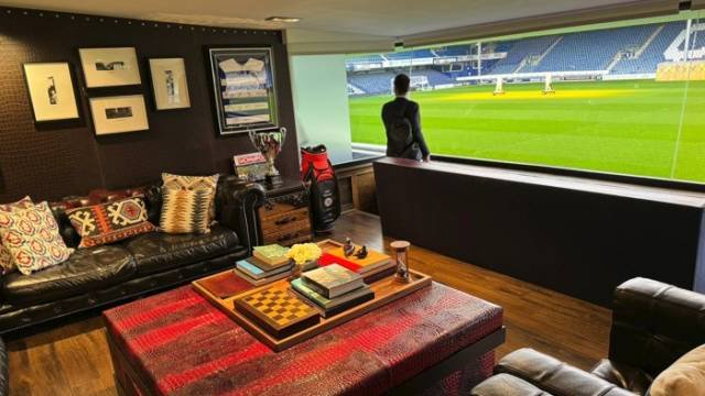Homem de terno escuro, de costas, observa o campo de futebol verde e as arquibancadas azuis de um estádio através de uma grande janela. À esquerda, uma sala luxuosa com sofá de couro, almofadas estampadas, mesa de centro vermelha com livros e tabuleiro de xadrez, e quadros na parede. Um ambiente sofisticado para assistir a jogos