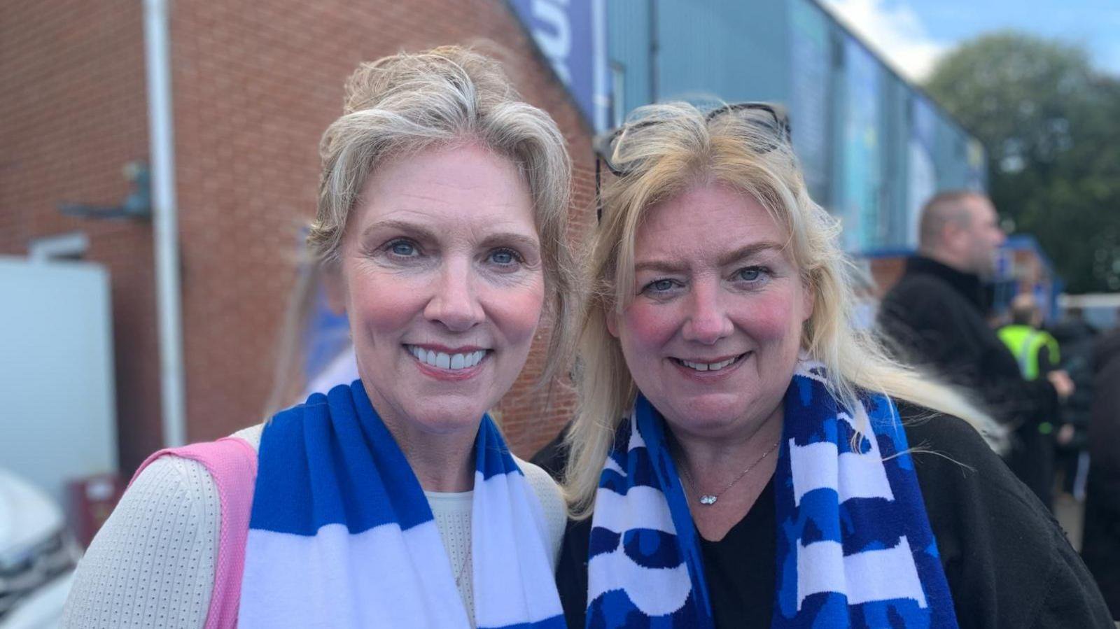 Tracy Grahams (left) and Lisa Hannaghan (right) wearing blue-and-white scarves pose for a photograph outside the football ground, with stadium buildings and other supporters visible behind them.