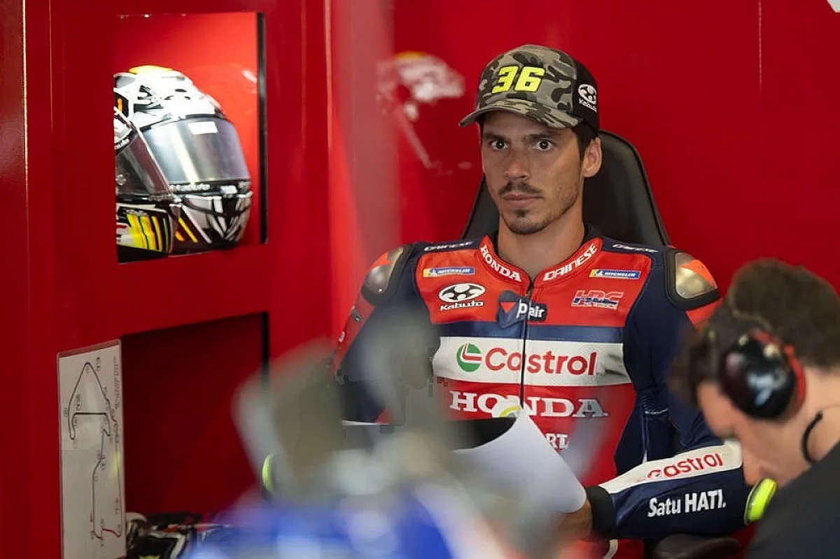 Rider in Honda HRC leathers with Castrol livery and a number 36 camo cap in the garage, MotoGP paddock setting, helmet on display