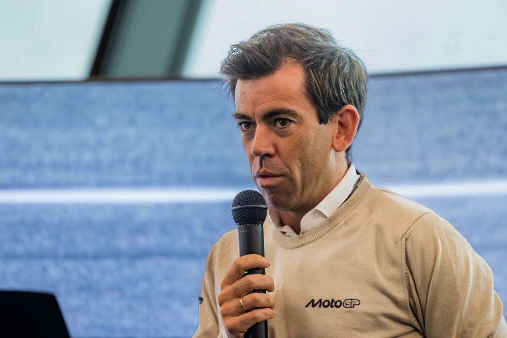 A man speaking into a microphone at a MotoGP event, wearing a beige sweater with the MotoGP logo, during a press conference setting.