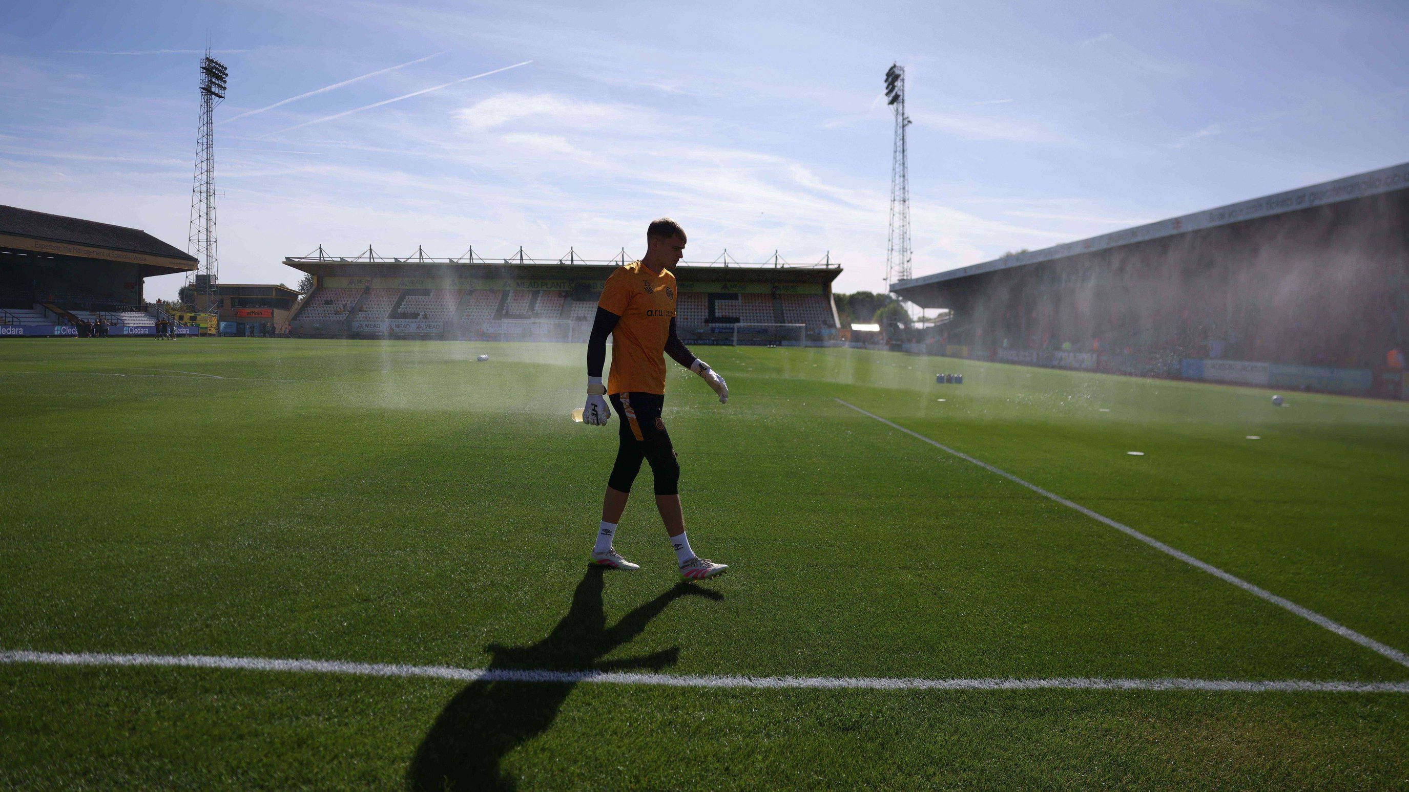 A man wearing a football kit and goalkeeping gloves walks across a green football pitch, which is surrounded by the four sides of a stadium. There is blue sky above.