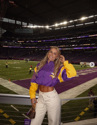 A woman in a purple and yellow Vikings jacket and white pants stands in a football stadium.