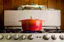 A red pot with a lid is on a vintage stove, with gas burners lit beneath it. The stove has an old-fashioned design with dials. No people are present