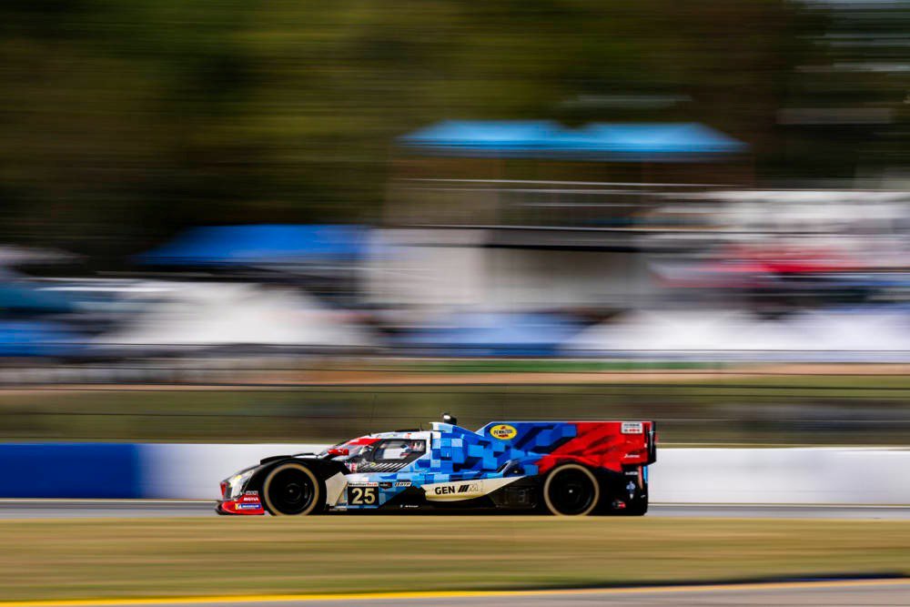 BMW M Hybrid V8 race car #25 in blue, red and white livery speeding on track at IMSA, with blurred pit buildings behind