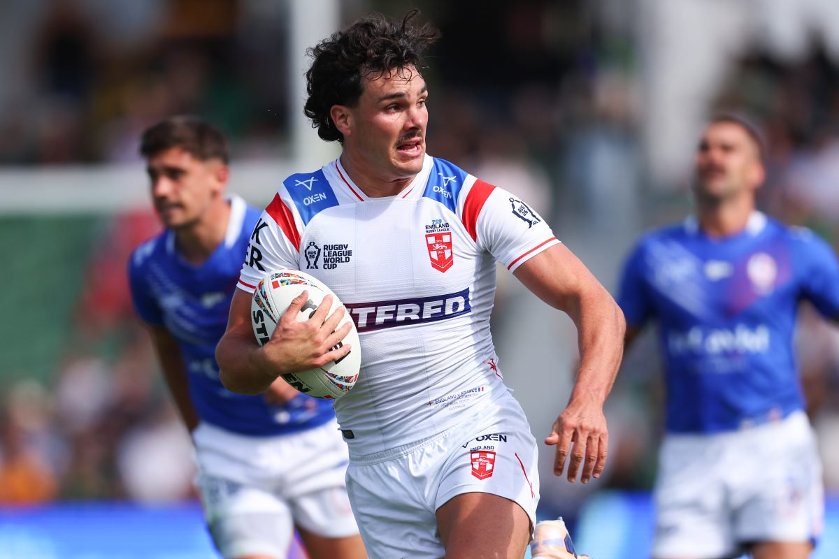 A male rugby player in a white England jersey runs with the ball on a sunny day.