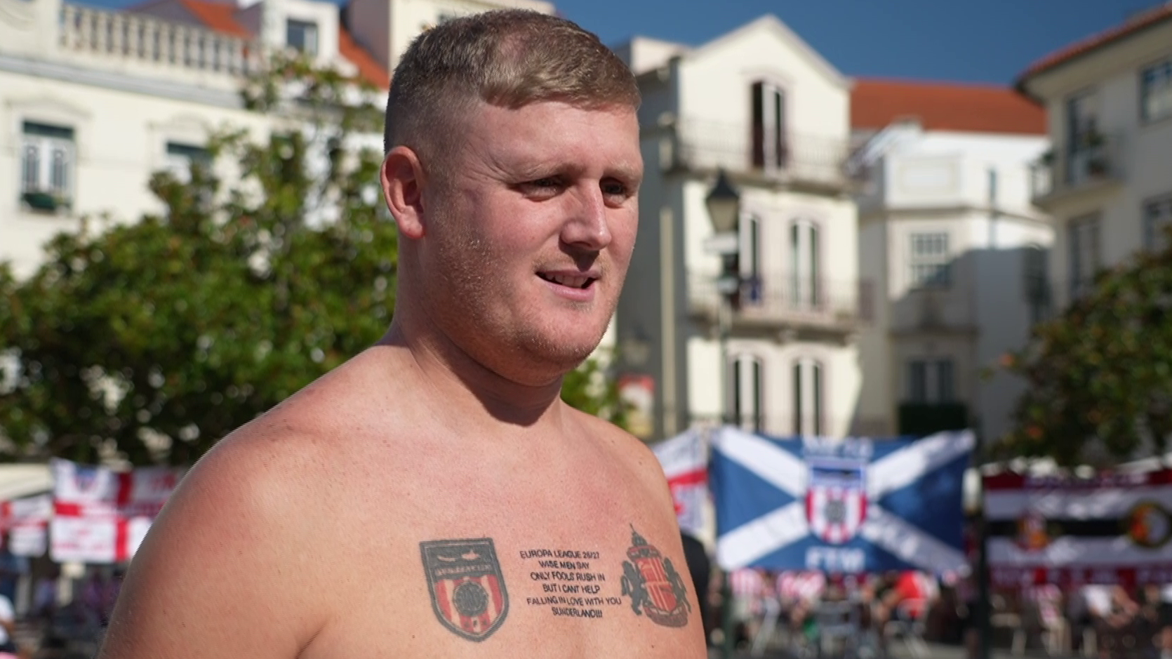Luke Thompson is topless and has light brown hair. He is looking at the camera and has a large chest tattoo which depicts the Sunderland AFC logo and the Torreense logo. In between the two logos there is writing. 