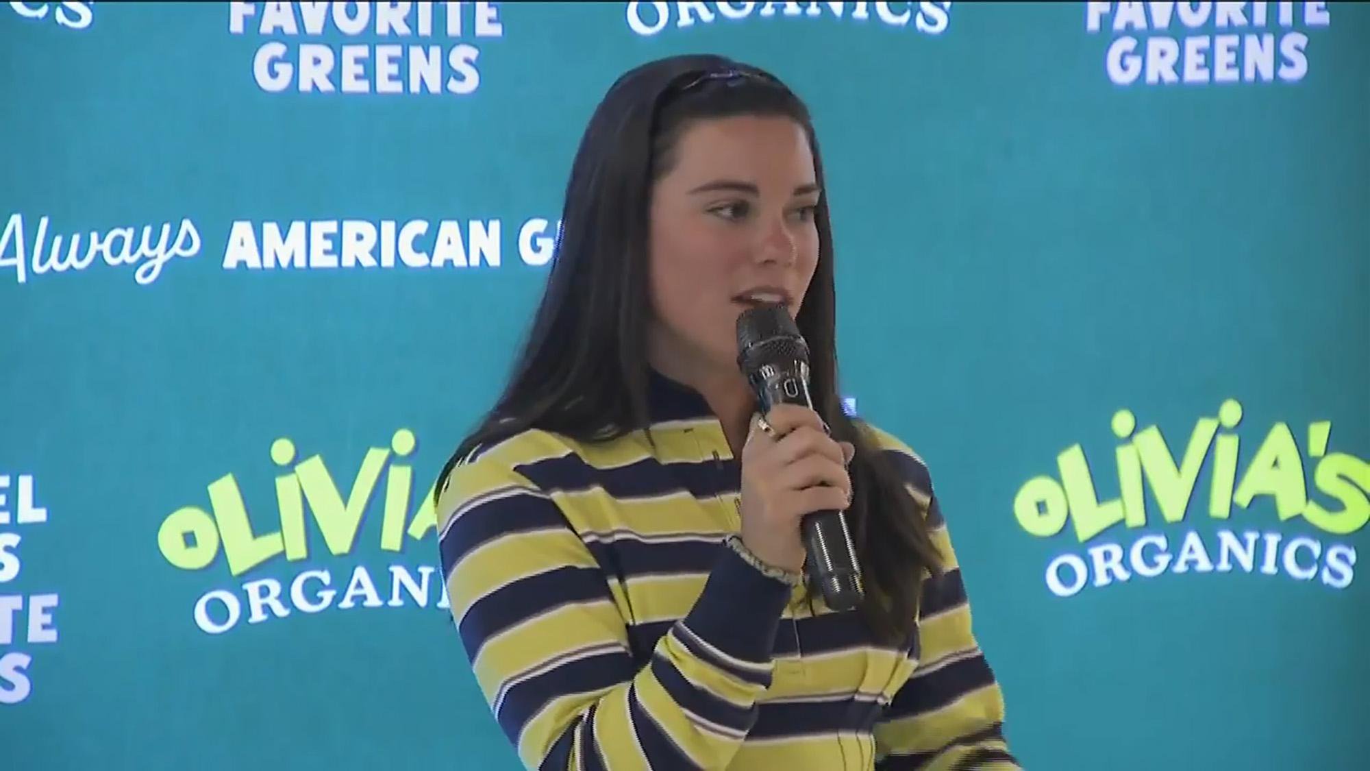 Ann Michael Maye speaking into a microphone in front of a branded backdrop.