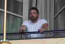 Ben Affleck stands on a balcony, leaning on the railing with a cigarette in his hand, looking at the distance