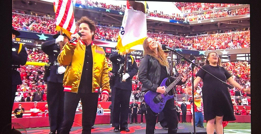 Pat Monahan, lead singer of Train, singing on a football field in front of a stadium crowd, accompanied by a woman playing electric guitar and an interpreter.