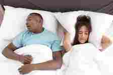 A man is sleeping on his back and snoring. A woman next to him covers her ears with a frustrated expression. Both are lying in bed under white blankets