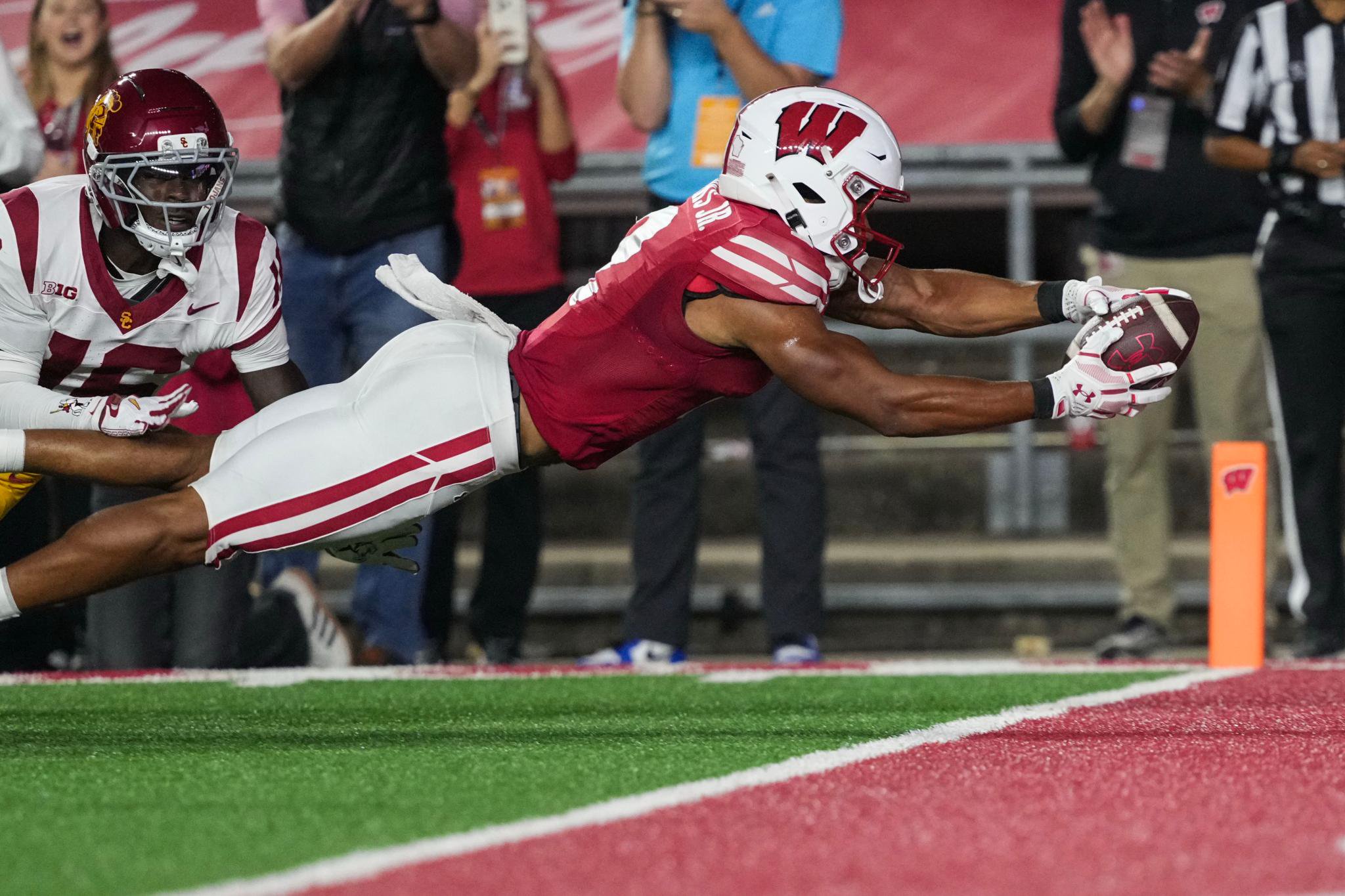 A Wisconsin football player dives to catch a football just outside the end zone while a USC defender pursues him.