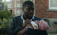 Kevin Hart holds a baby wrapped in a blanket, standing outside with buildings in the background