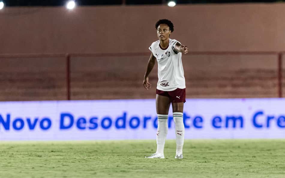 Anny, zagueira, em campo durante o jogo, com uniforme branco e meiões brancos, diante de publicidade azul no estádio.