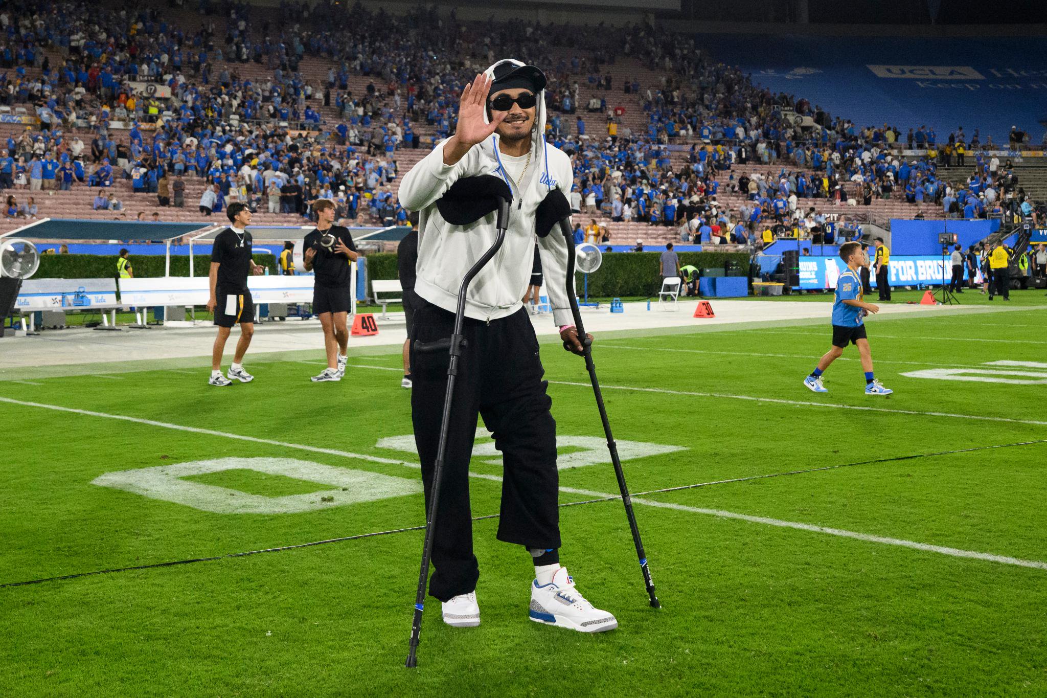 Nico Iamaleava on crutches, smiling and waving to the crowd on the football field.