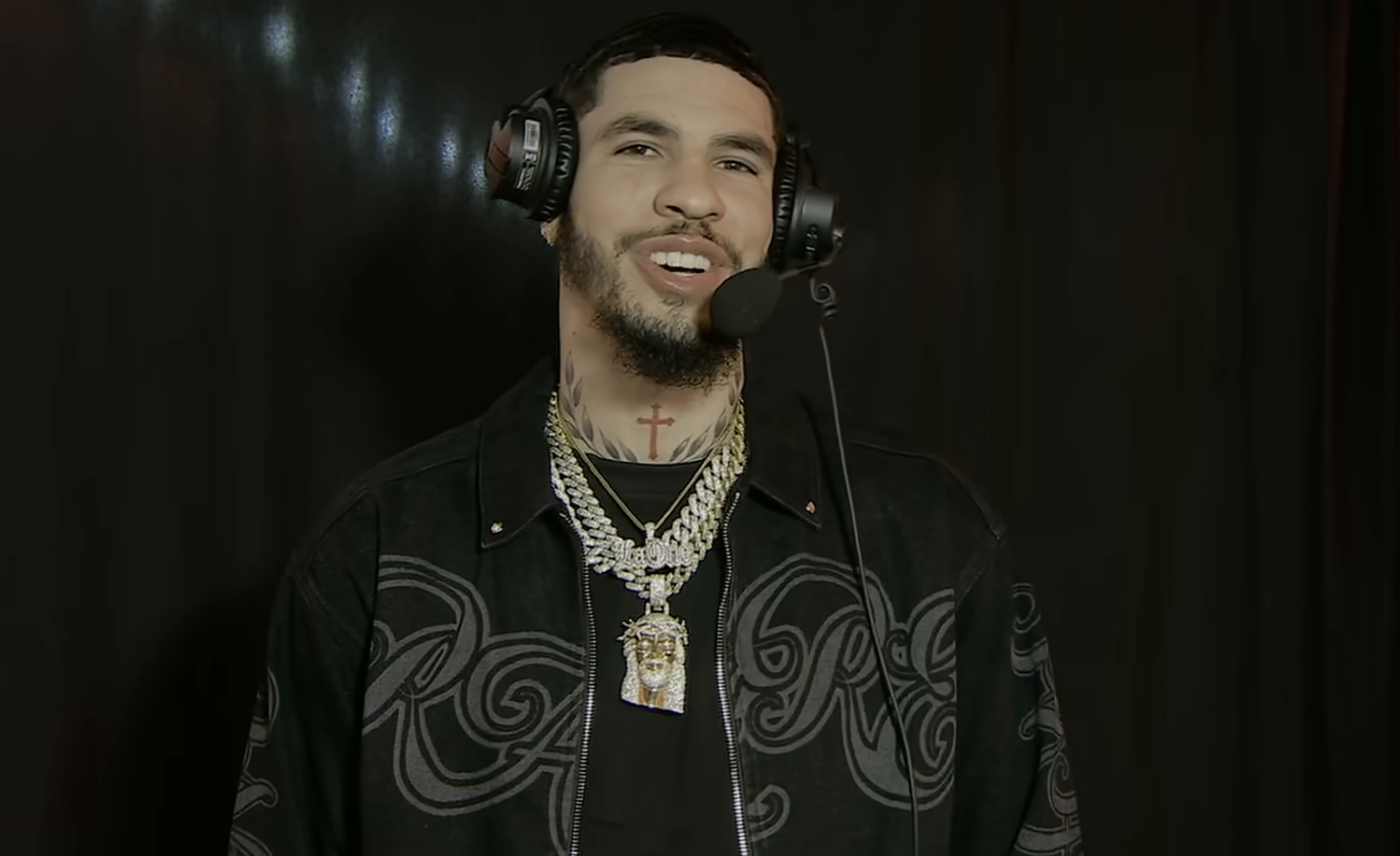 A person is speaking into a headset microphone, wearing a patterned jacket and several layered necklaces