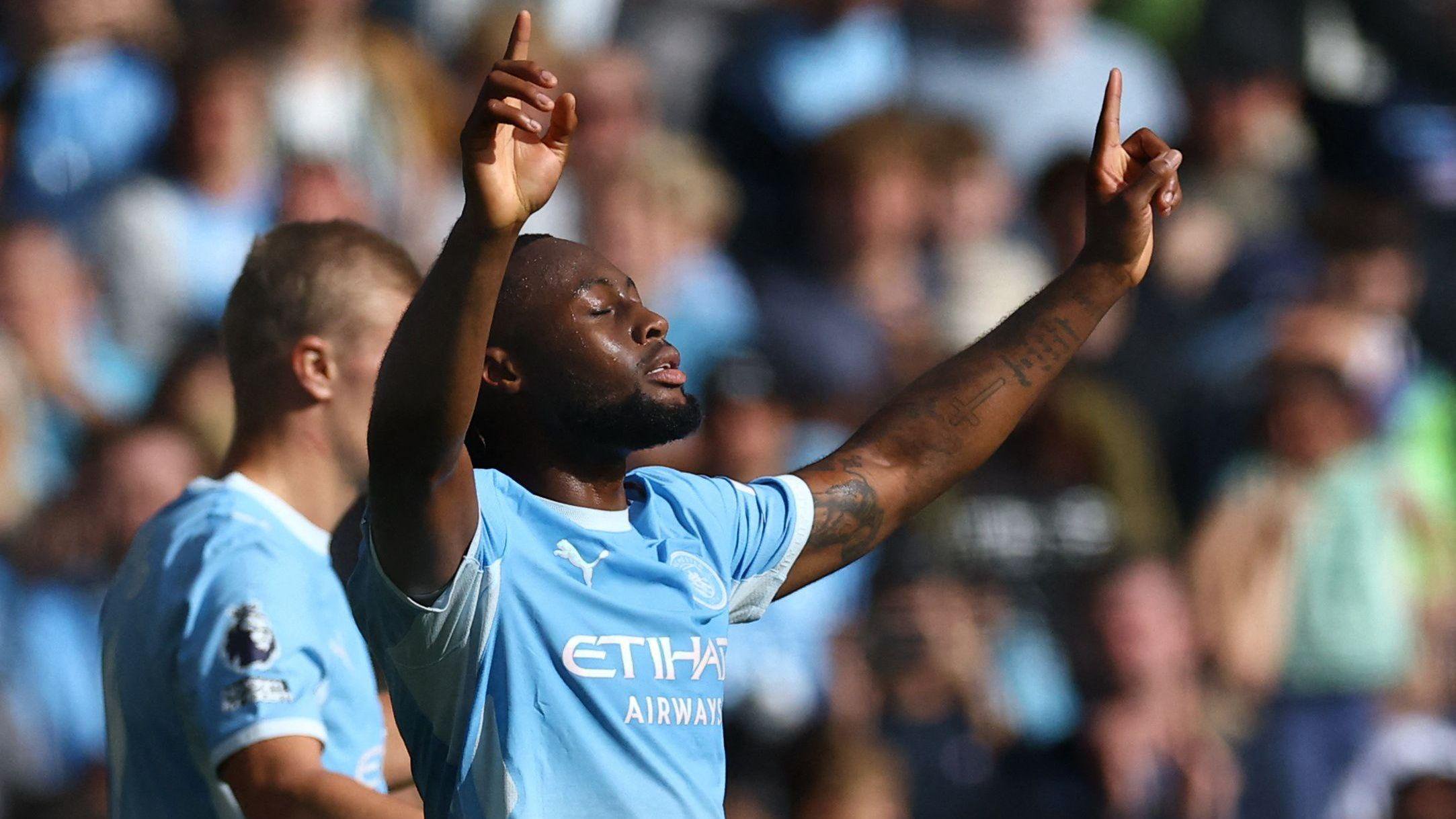 Semenyo wearing Manchester City's sky-blue home shirt stands on the pitch with both arms raised above his head and his index fingers pointing upwards. His eyes are closed. A large crowd of spectators fills the blurred background.