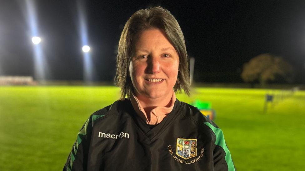 Nia Evans is photographed standing in front of a rugby pitch. She is wearing a black and green rugby jersey. She has dark grey shoulder length hair. She is smiling and looking at the camera. 
