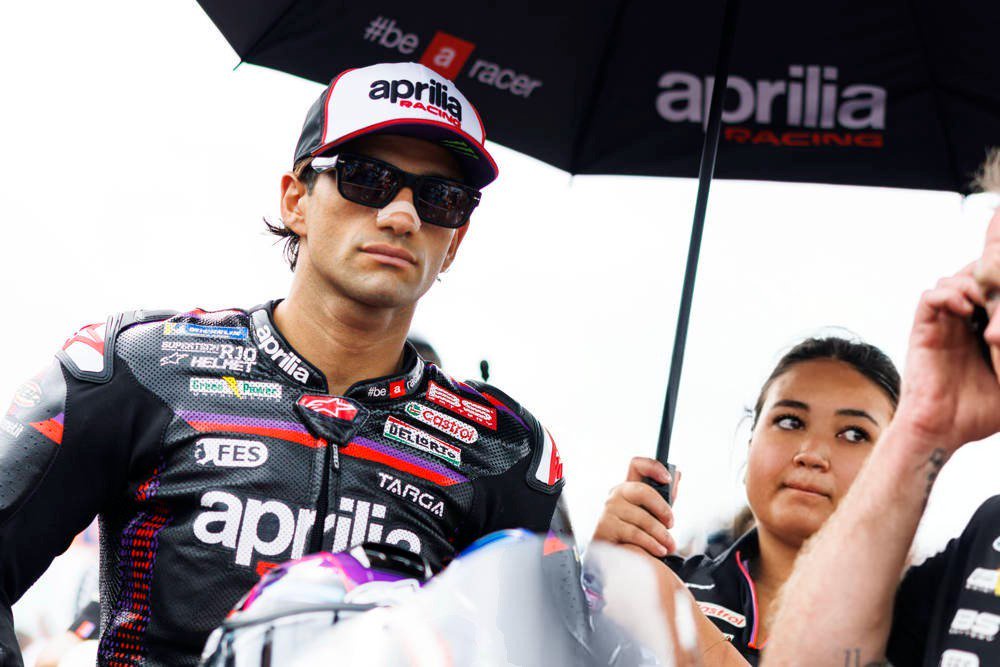 Rider in black Aprilia Racing leathers and cap under a team umbrella in the MotoGP paddock, with a woman standing nearby