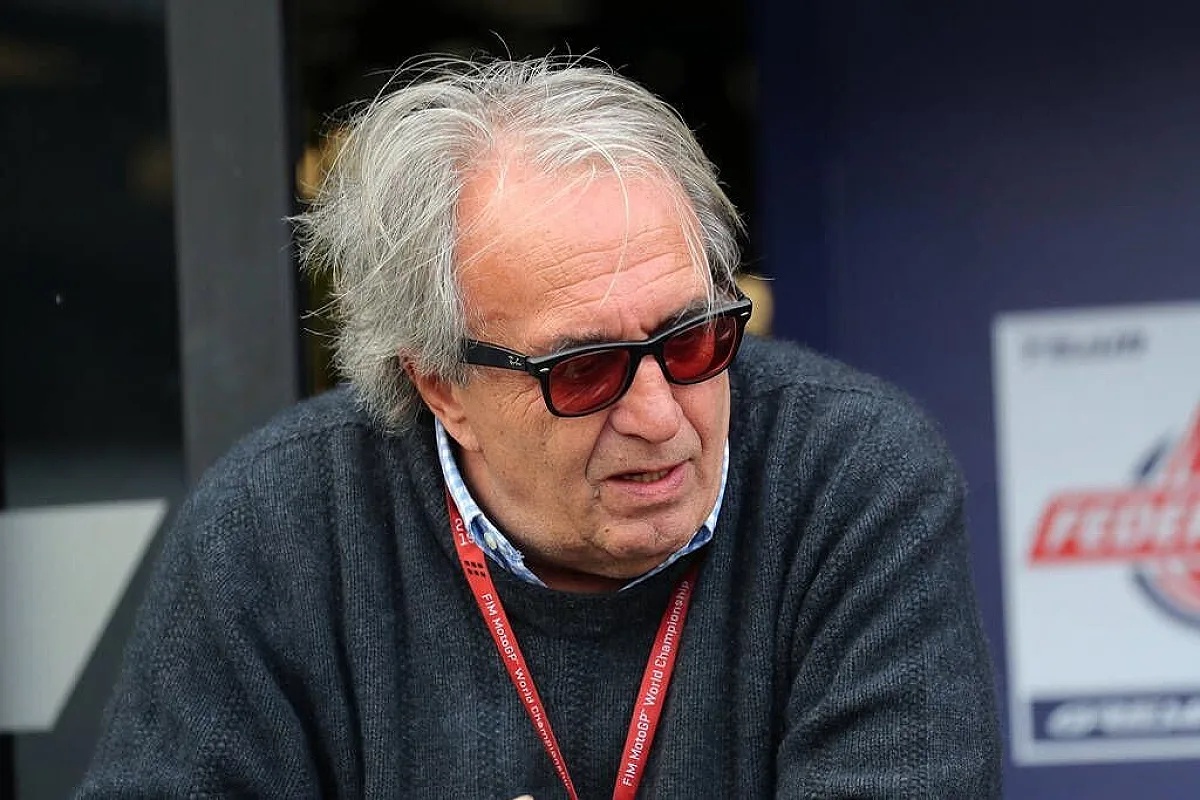 A man with grey hair and tinted red sunglasses, wearing a dark sweater and a FIM MotoGP World Championship lanyard, sits near a Federal Oil banner in the pad…