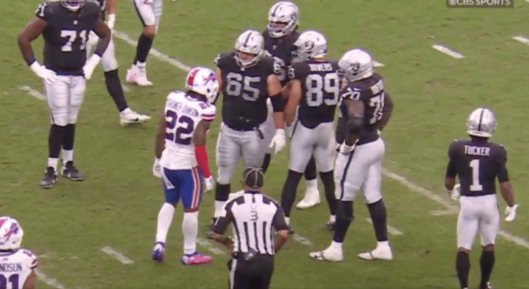 A football player in a white and blue uniform faces a group of opposing players in black and silver uniforms, while a referee stands nearby.