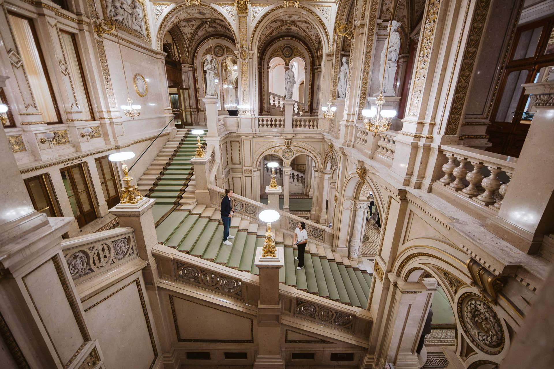 Zwei Personen stehen auf einer prunkvollen, breiten Marmortreppe in einem historischen Gebäude.