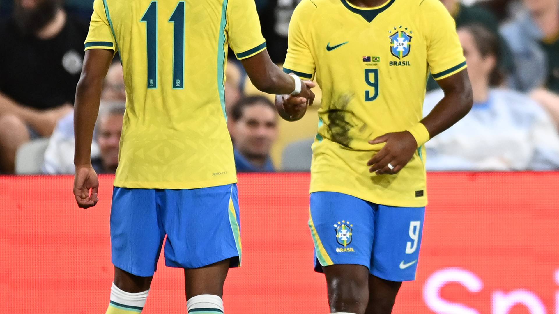 Dois jogadores de futebol brasileiros, Estêvão (camisa 11) e Rodrygo (camisa 9), apertam as mãos no campo, sorrindo. Ambos vestem uniformes amarelos e azuis, com meias brancas e detalhes coloridos. Torcedores desfocados aparecem ao fundo.