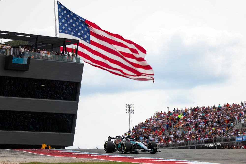 Kimi Antonelli's Mercedes F1 car speeds past grandstands and a US flag at the 2026 United States Grand Prix