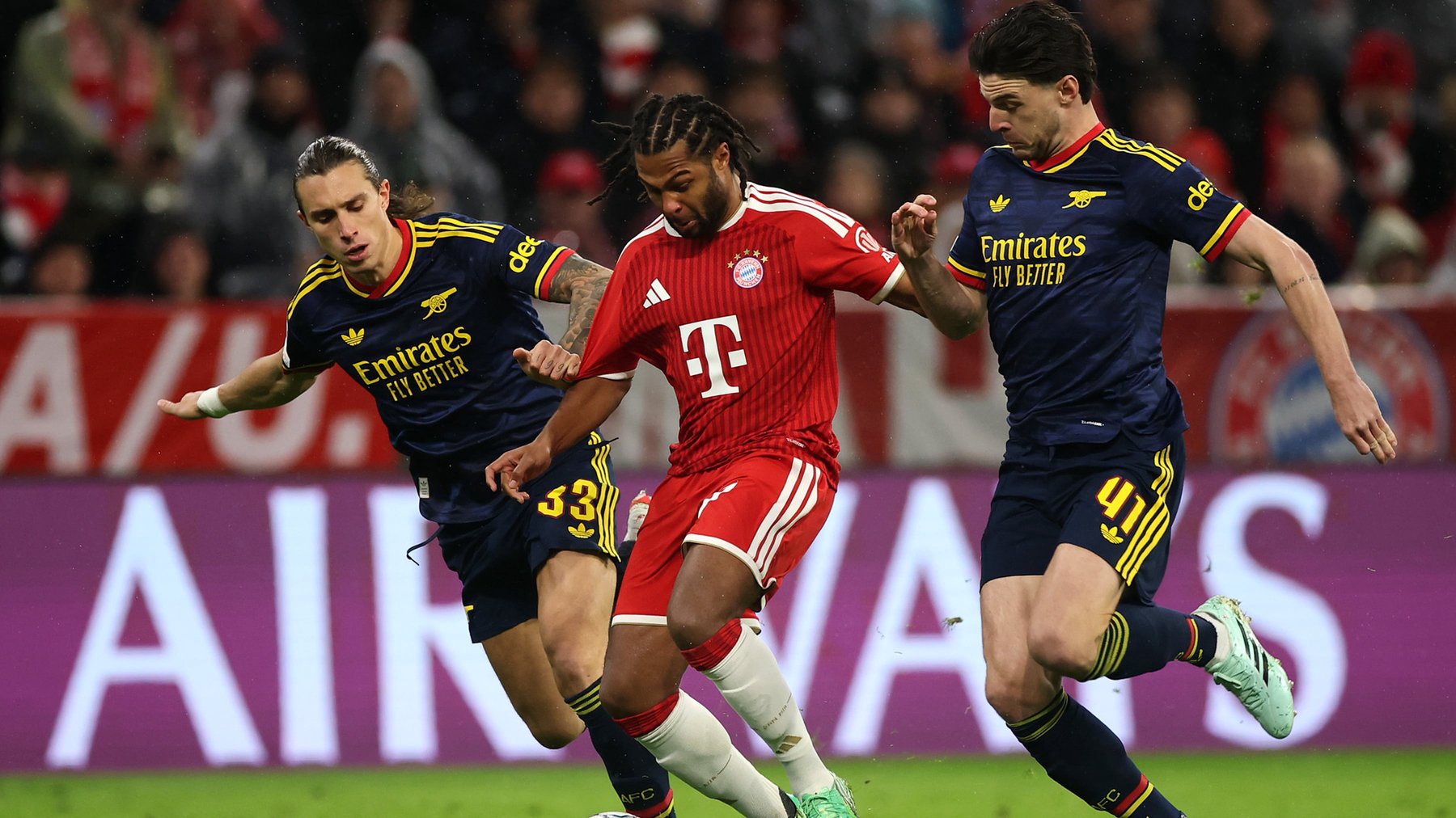 MUNICH, GERMANY - OCTOBER 21: Serge Gnabry of FC Bayern Munich runs with the ball under pressure from Riccardo Calafiori and Declan Rice of Arsenal during the UEFA Champions League 2026/27 League Phase MD3 match between FC Bayern München and Arsenal FC at