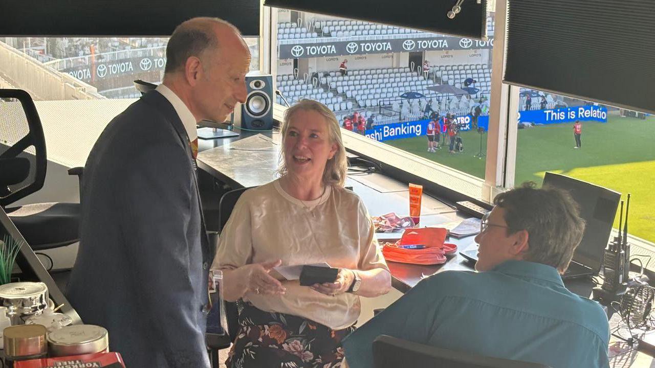 A man in a suit stood next to Jane and another person who are sat down in a cricket scoring box. Jane is holding a box with a medal in.