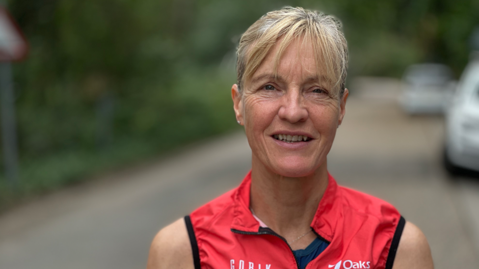 Catherine Linney stands on a road wearing a red running vest. There are parked cars and greenery lining the road behind her. She smiles at the camera.