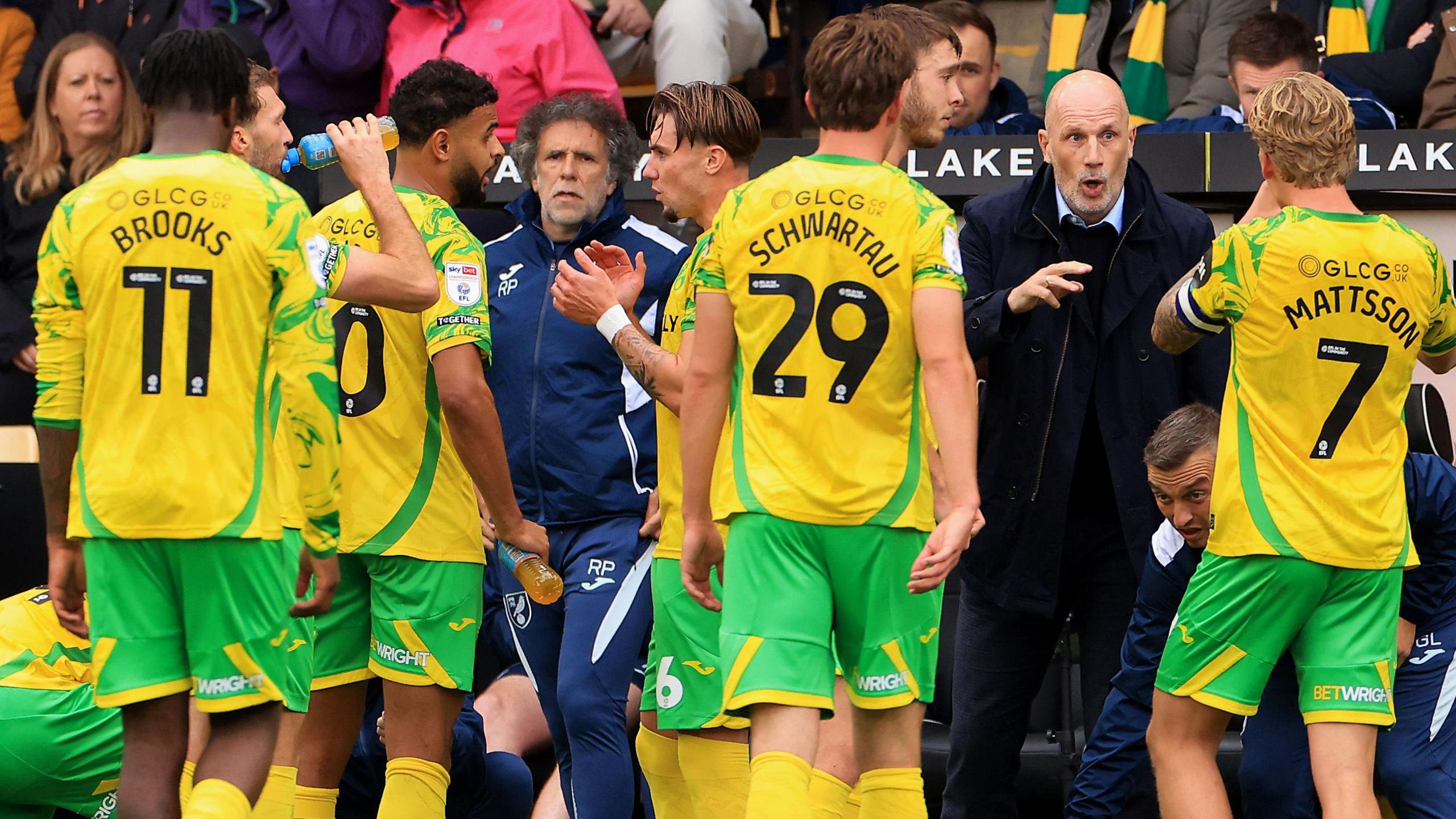 Philippe Clement, wearing a navy blue jacket, navy blue jumper and light blue smart shirt talks to his Norwich players, who are wearing their yellow and green home kit, on the sidelines during a game against Portsmouth at Carrow Road