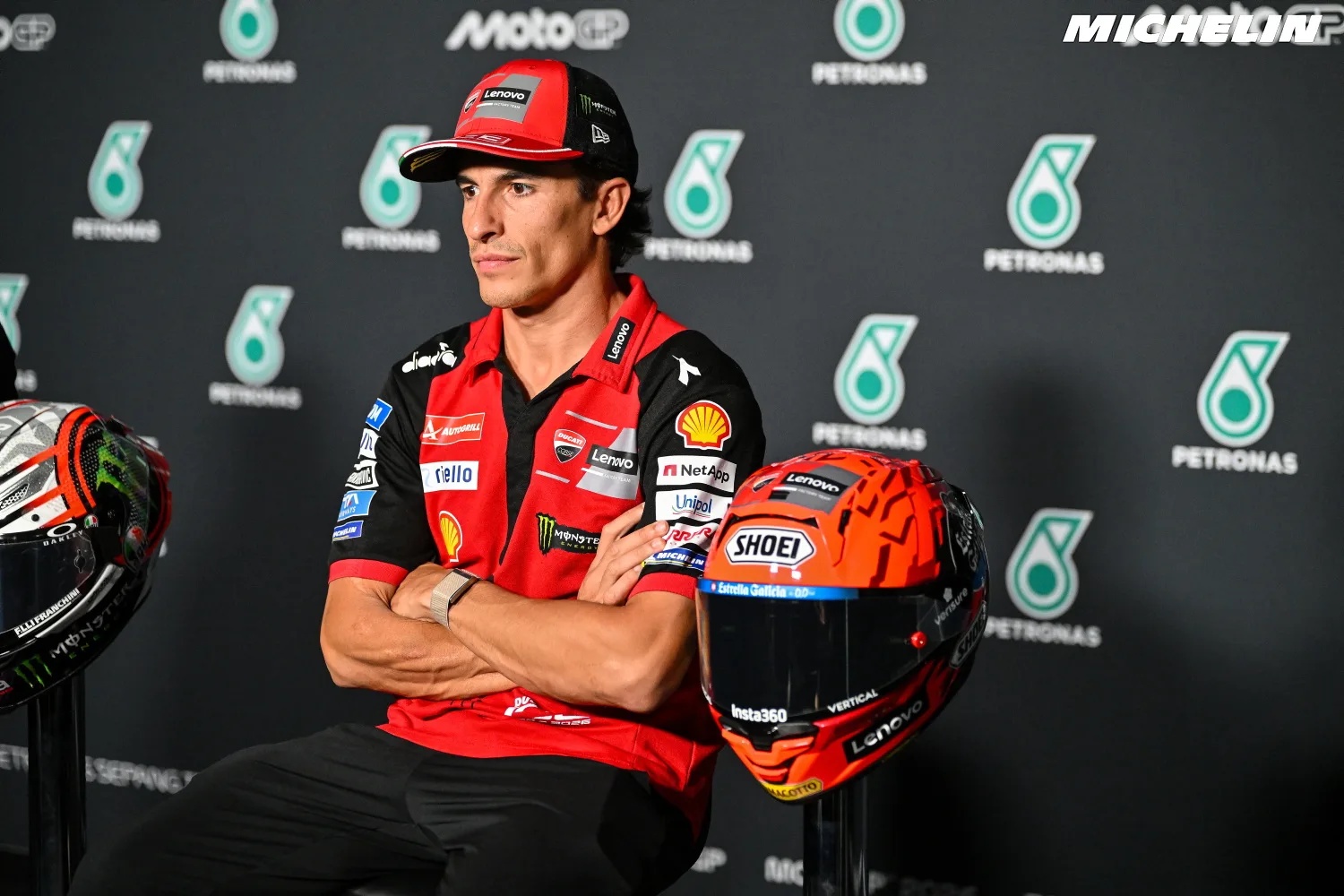 A Ducati rider in a red and black team shirt and cap sits arms crossed at a MotoGP press conference, with an orange helmet beside him.