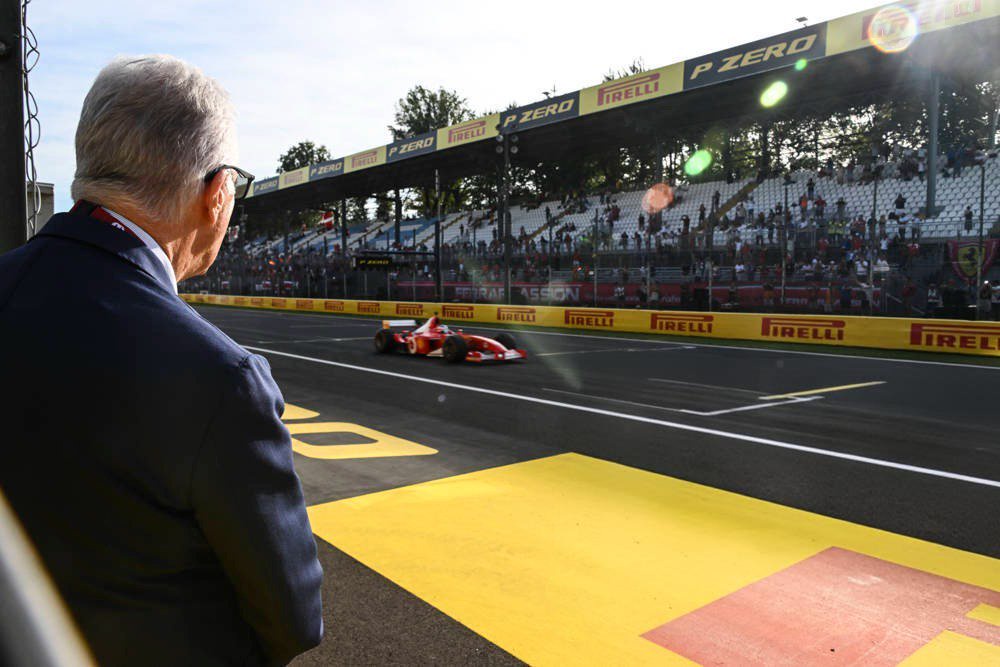 A man with grey hair in a navy jacket watches a red Ferrari F1 car on track at Monza, Pirelli-lined barriers in view.
