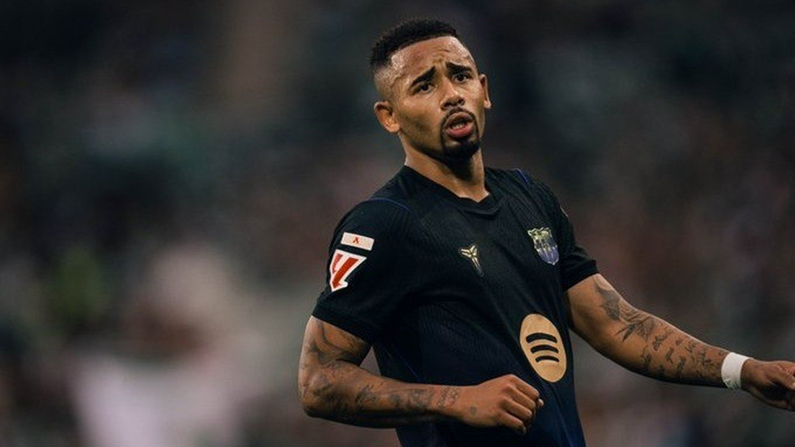Gabriel Jesus em campo, camisa escura de futebol, barba, tatuagens, boca semiaberta