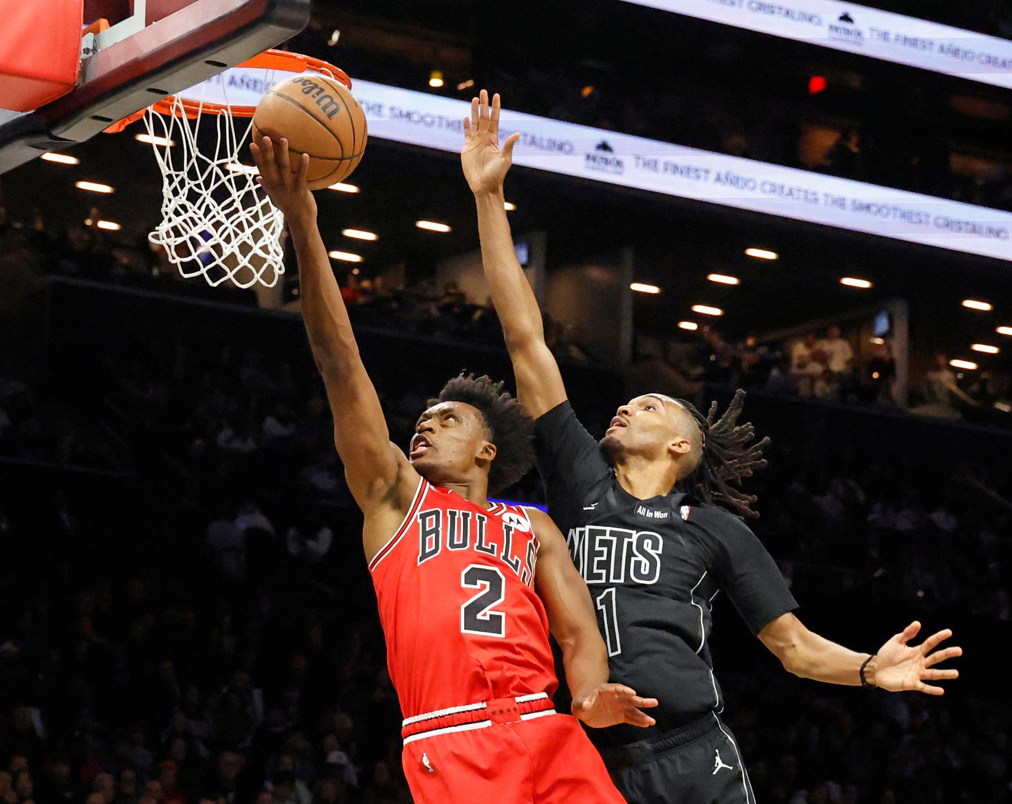 A Chicago Bulls player in a red jersey shoots a layup as a Brooklyn Nets defender contests the shot near the basket.