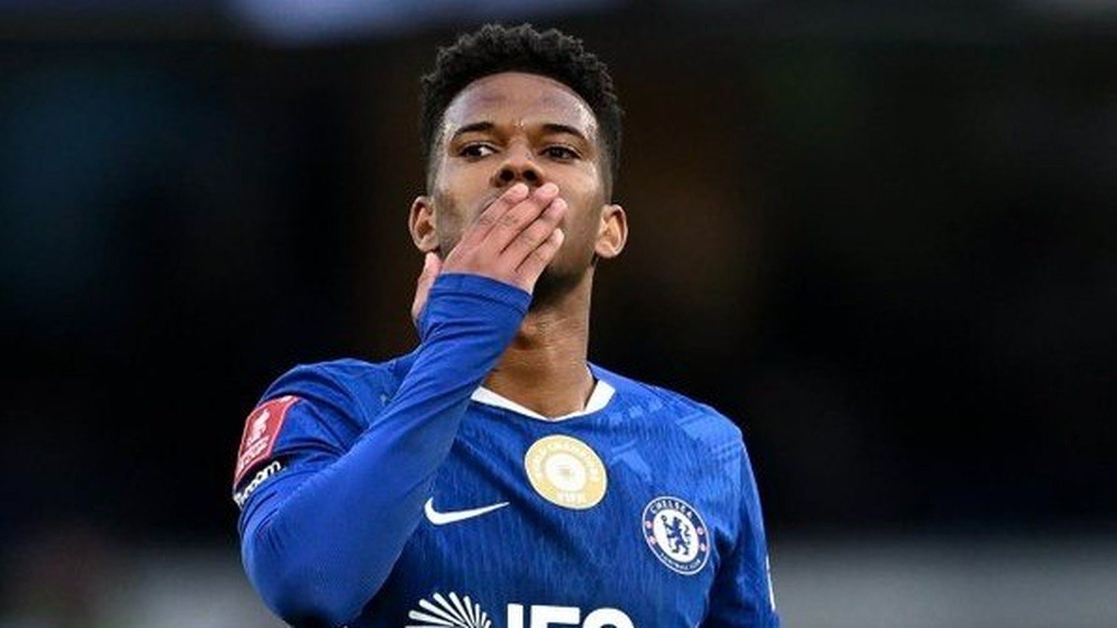 Jovem jogador de futebol, Estêvão, em camisa azul do Chelsea, soprando um beijo em campo