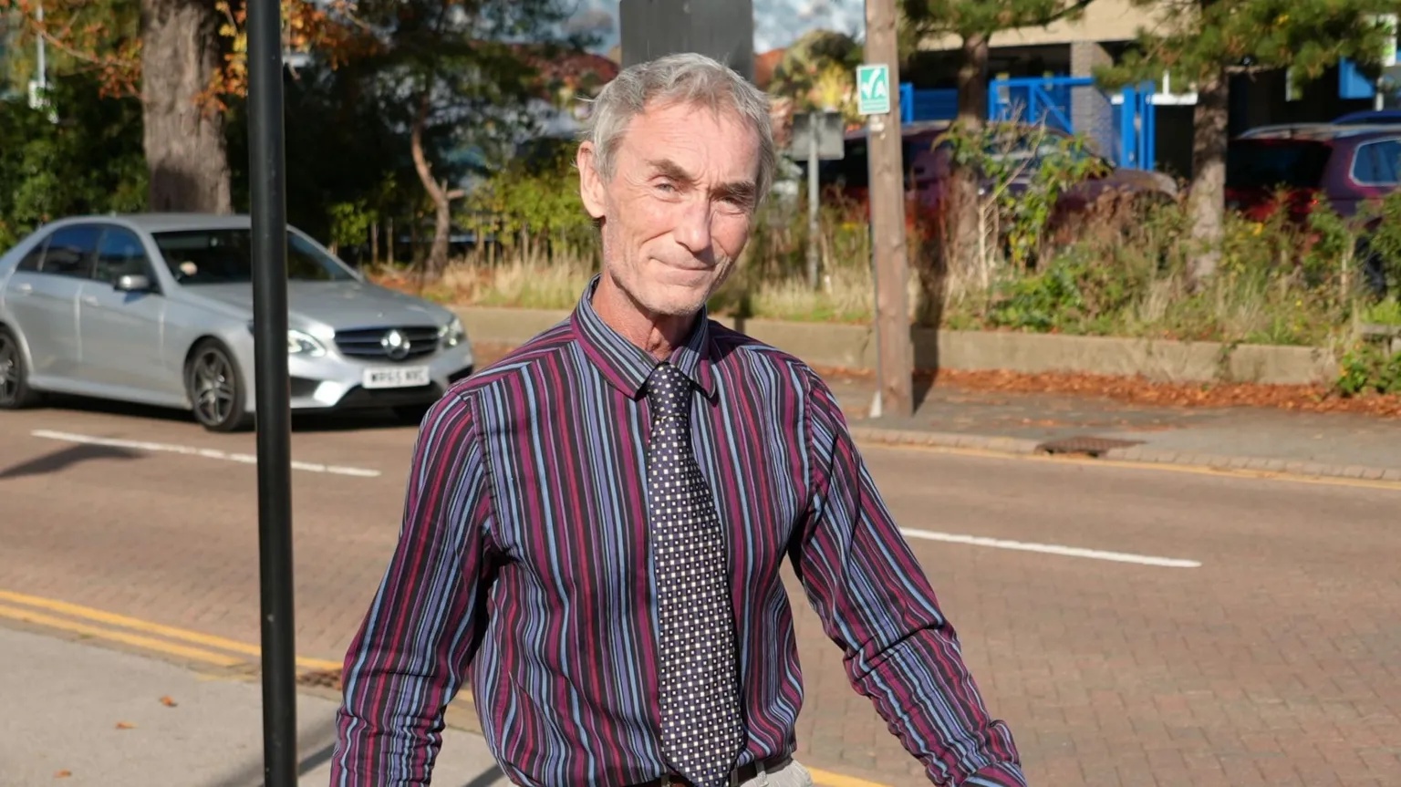 Shaun Whitmore/BBC A man smiles at the camera as he walks along a road. He wears a red, blue and purple striped shirt and a polka dot tie. He has short grey hair. 