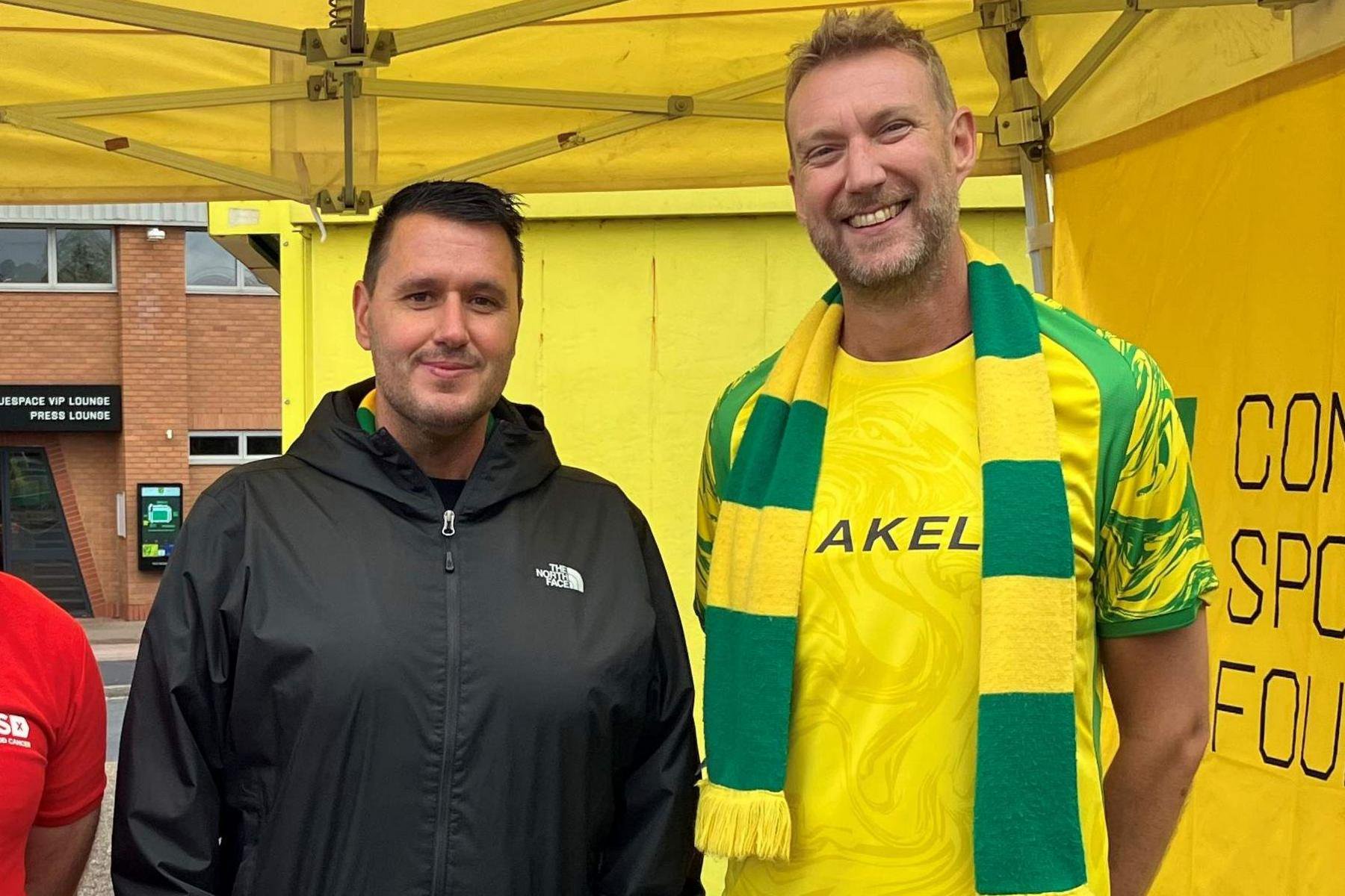 Two men stand next to each other. The one on the left has black short hair and is wearing black rain jacket on with a white logo on the top right of it. The man on the right is wearing a yellow and green Norwich City Football Club T-shirt. He is also wearing the club scarf. He has short light blond hair and a blond beard. Both the men are smiling at the camera. The man on the left has a closed smile while the man on the right is showing his teeth. Behind them is a bright yellow canopy, which they are standing under. 