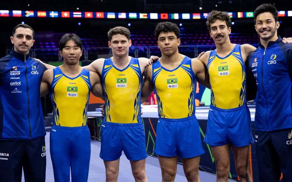 Seis ginastas brasileiros em amarelo e azul, com dois técnicos, posam em arena no Mundial de ginástica artística.