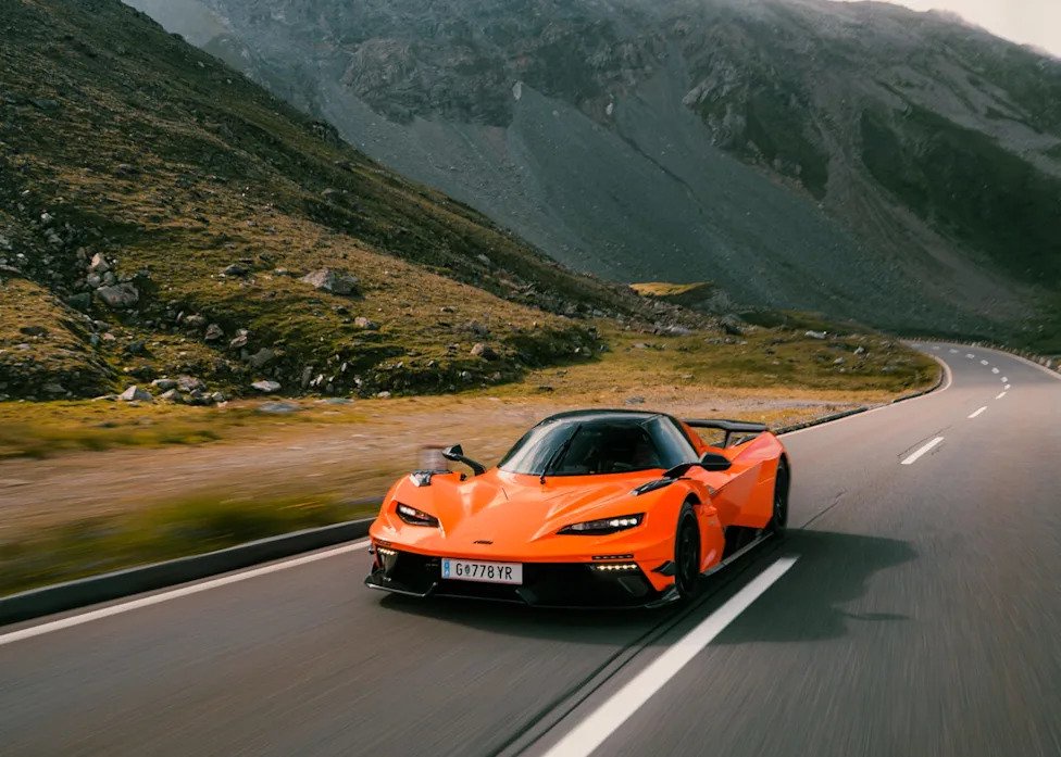 Bright orange sports car driving on a winding road through a mountainous landscape.