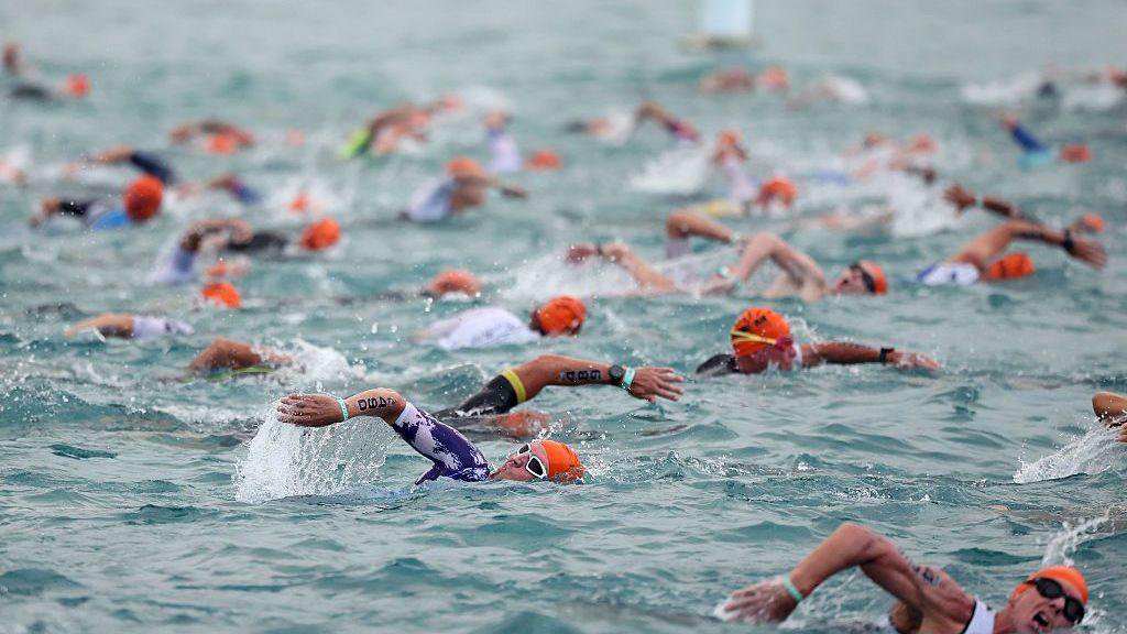 Athletes in orange swim caps compete in the Ironman World Championship in Kailua-Kona, Hawaii, on 10 October. Dozens of competitors are swimming in the sea while wearing wetsuits and swimming goggles. They are all performing the front crawl with their arms stretched out. The water looks relatively still, with ripples being caused by the athletes' swimming strokes.