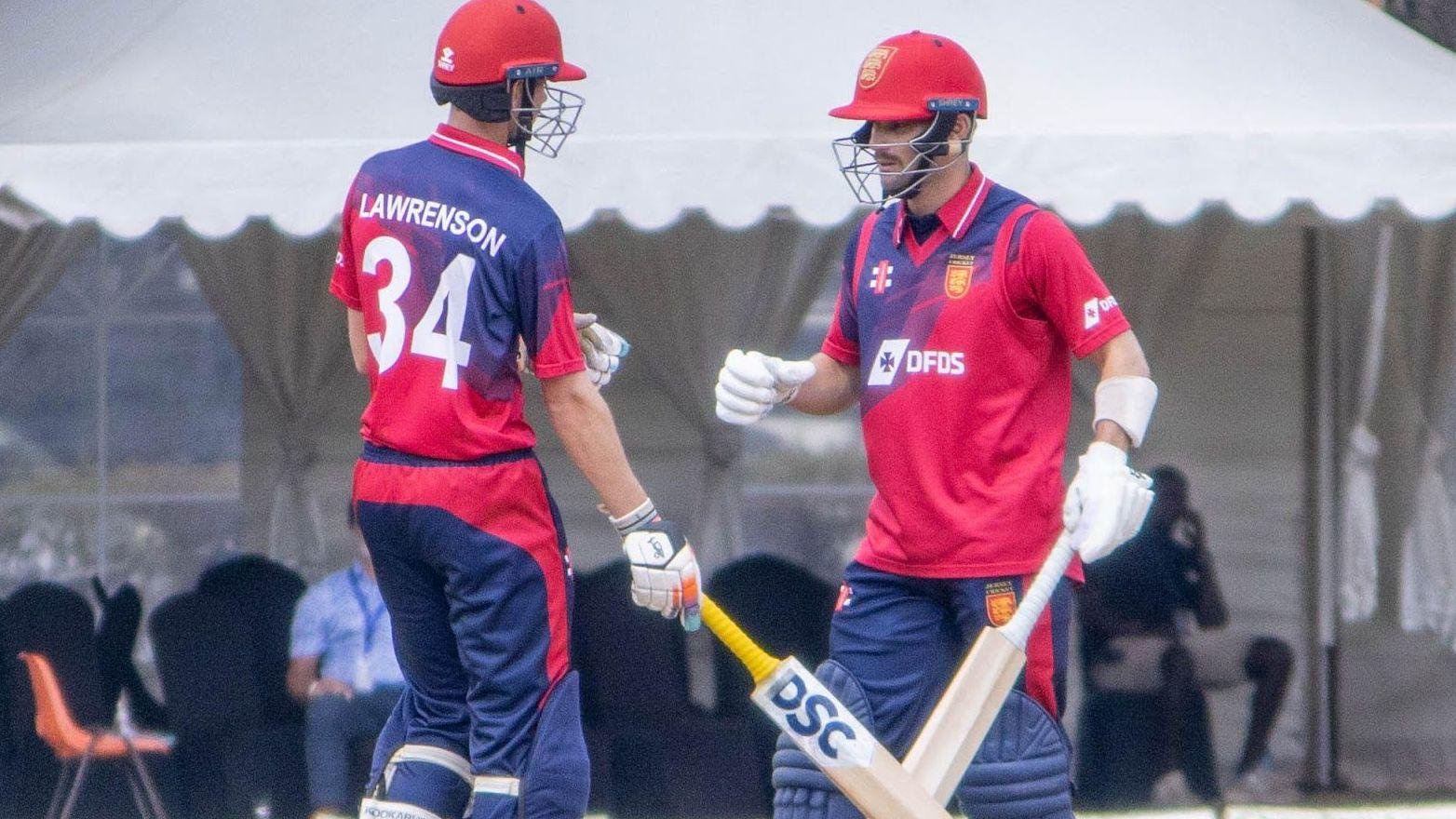 Josh Lawrenson and Jonty Jenner wear red helmets, red-and-blue jerseys, and white gloves while holding cricket bats. A pavilion and a row of dark-coloured chairs are in the background.